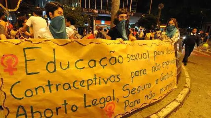 Manifestantes com o rosto coberto seguram, à noite, uma faixa sobre educação sexual, contraceptivos e aborto legal